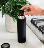 Person using a black coffee grinder with a plant and kitchen items in the background