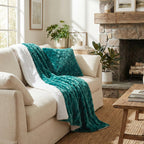 A teal textured throw blanket and a matching pillow are draped over a cream sofa in a bright, sunlit living room with a stone fireplace, plants, and wooden furniture.